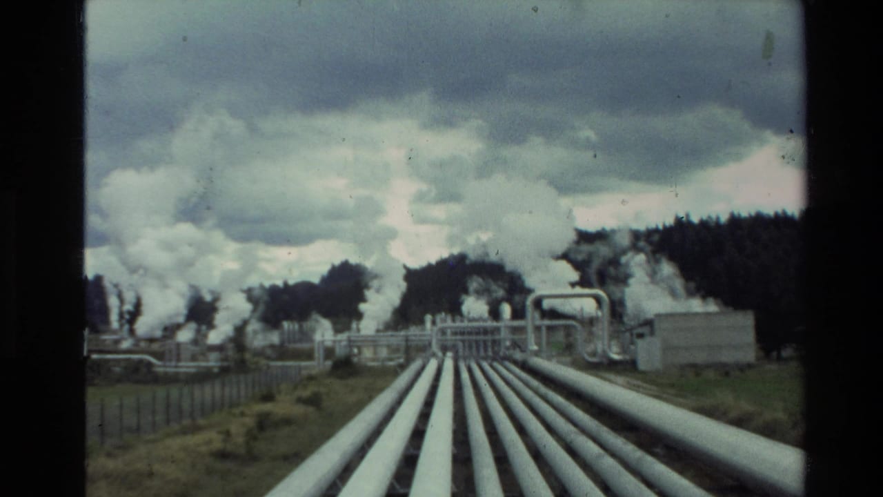 Wide 8mm frame of a New Zealand geothermal facility, with pipe runs crossing grass toward steam-emitting structures.