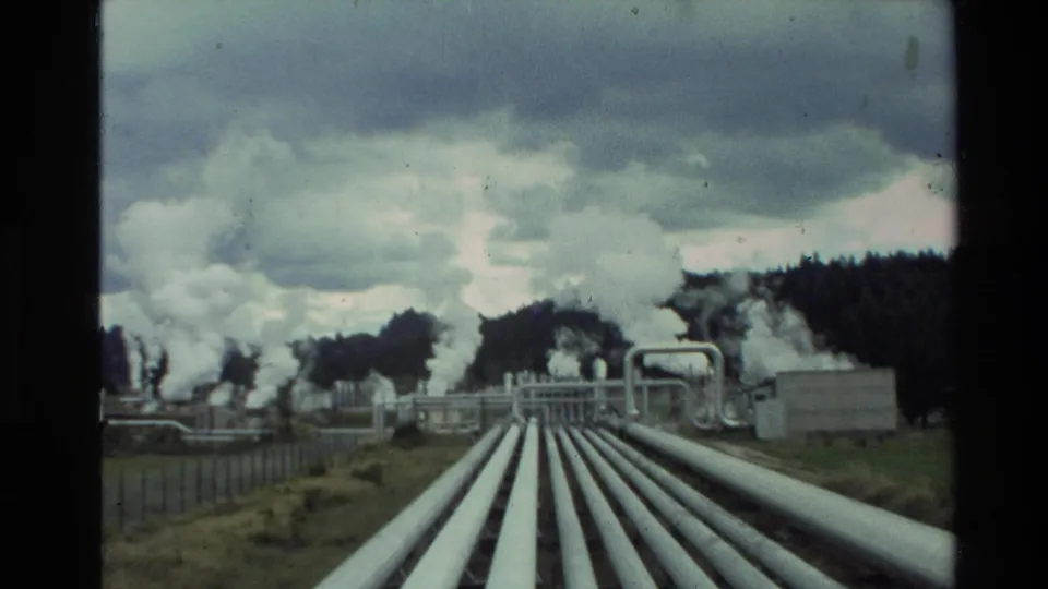 Wide 8mm frame of a New Zealand geothermal facility, with pipe runs crossing grass toward steam-emitting structures.