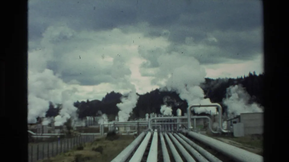 1981 New Zealand home-movie frame looking along geothermal pipes toward a fenced plant and rising white steam.