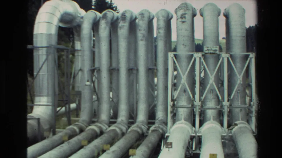 Wider 1981 New Zealand 8mm view of clustered insulated pipes and metal support frames near a wooded landscape.