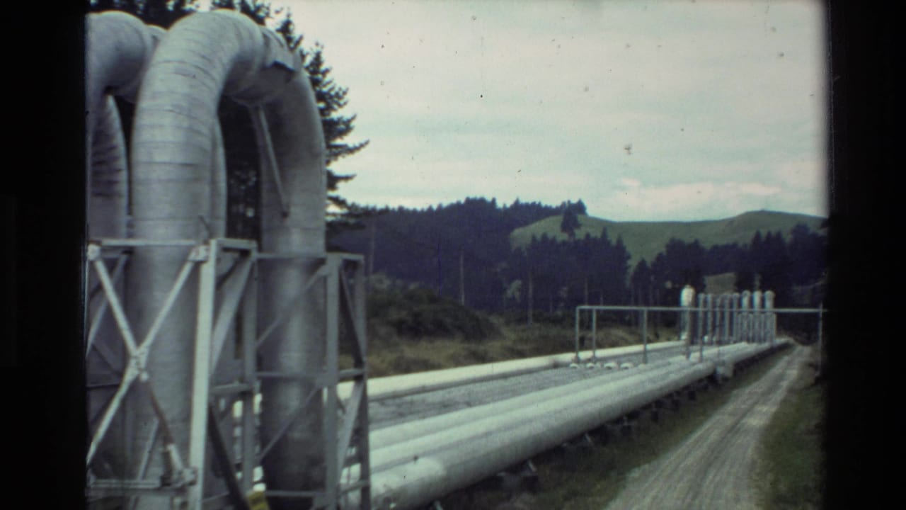 1981 New Zealand 8mm frame showing industrial pipework on supports beside a dirt road, with trees and low hills in the distance.