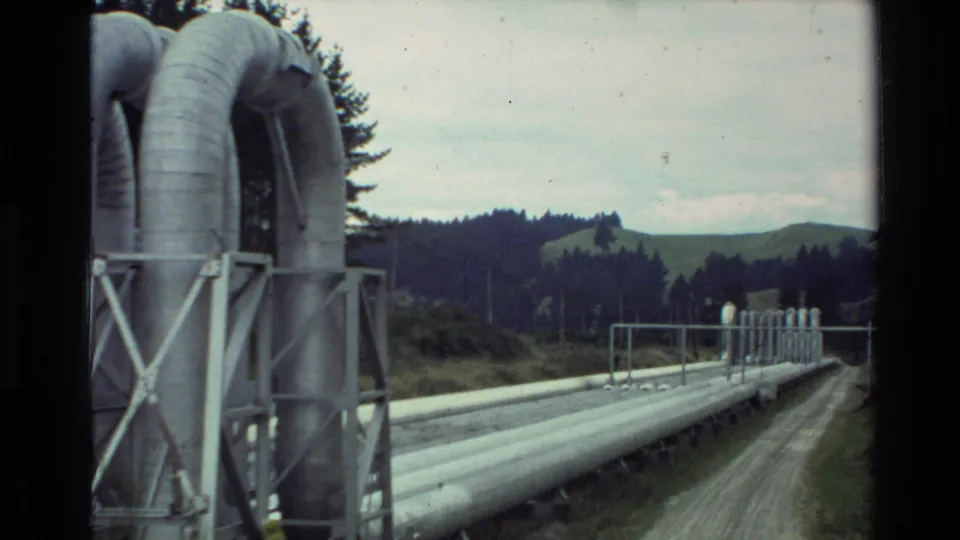 1981 New Zealand 8mm frame showing industrial pipework on supports beside a dirt road, with trees and low hills in the distance.