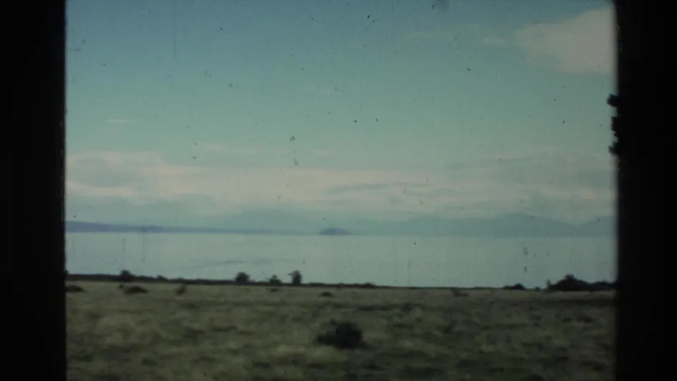 1981 New Zealand home-movie frame of an open plain, still water, faint hills, and a small distant island or headland through a dark frame.
