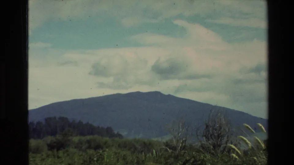 1981 New Zealand home-movie frame of a broad mountain ridge, green hills, and cloudy sky with faded 8mm color.