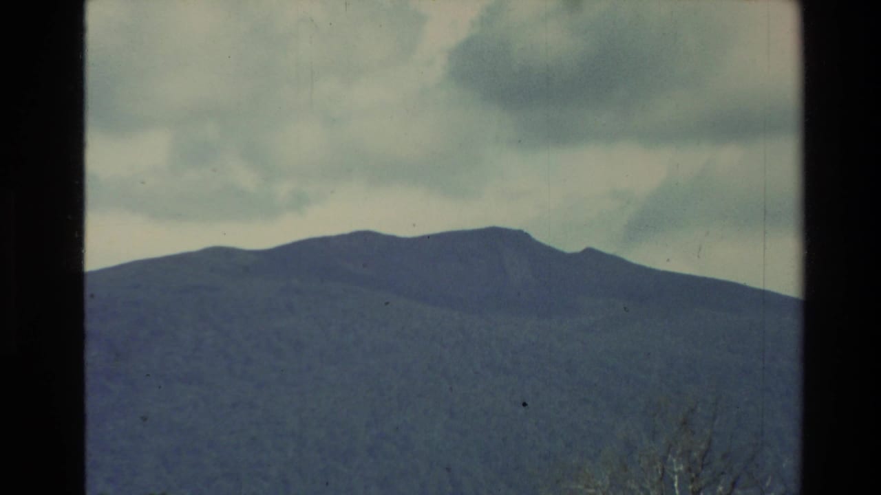 Broad blue-green mountain range below large clouds, framed by dark film borders.