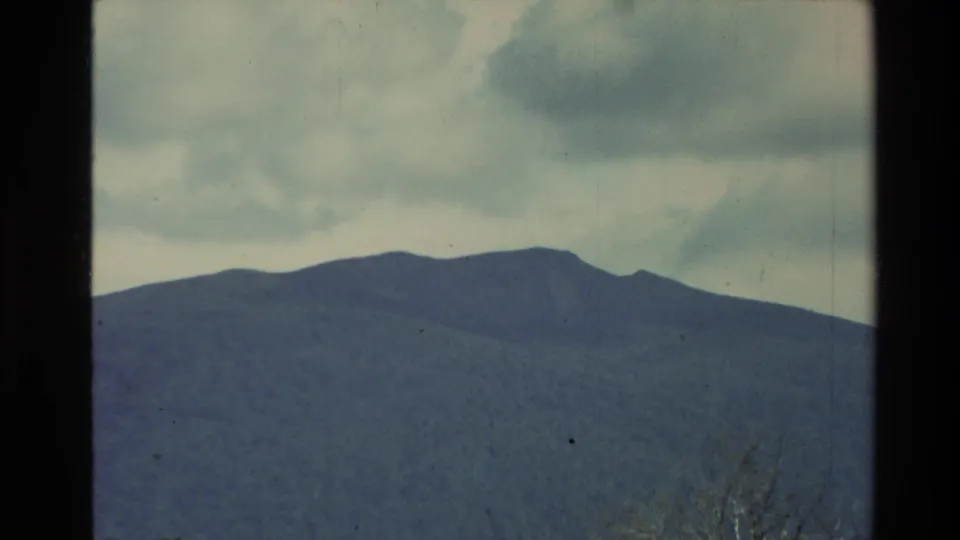 1981 New Zealand 8mm frame with a broad mountain range and subdued forested slopes beneath large, dark clouds.