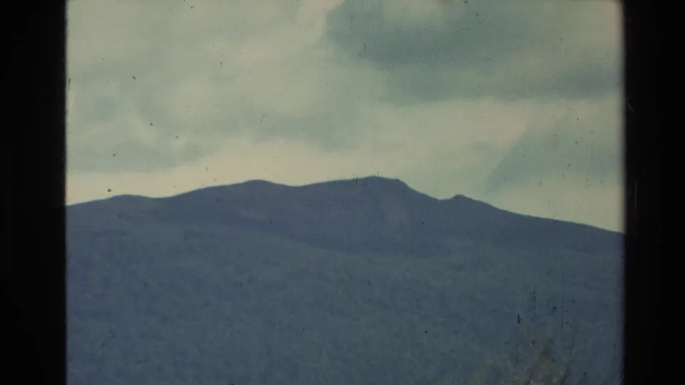 1981 New Zealand 8mm frame showing rounded mountain ridges and dark wooded slopes under a pale, cloud-filled sky.