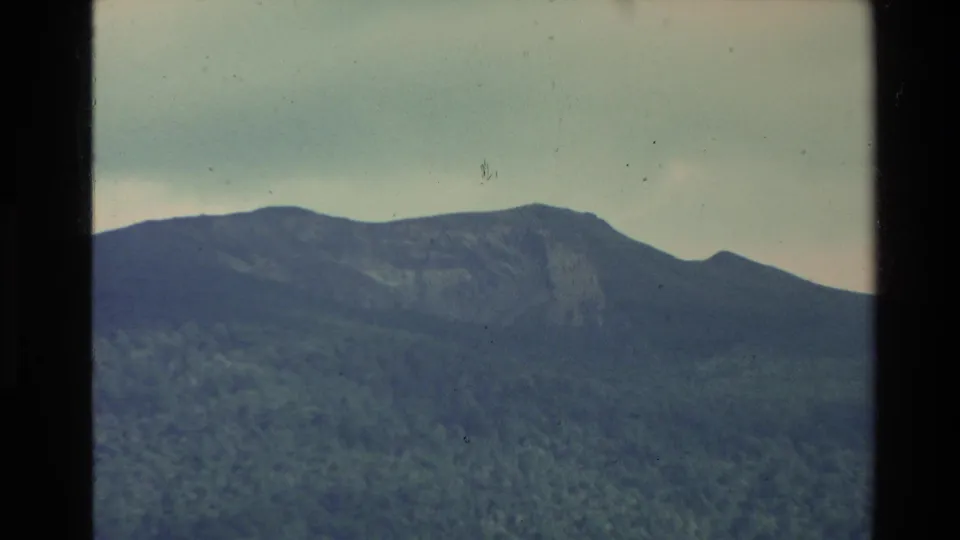 1981 New Zealand 8mm frame showing a darker forested mountain face with cloud cover overhead and visible home-movie film texture.