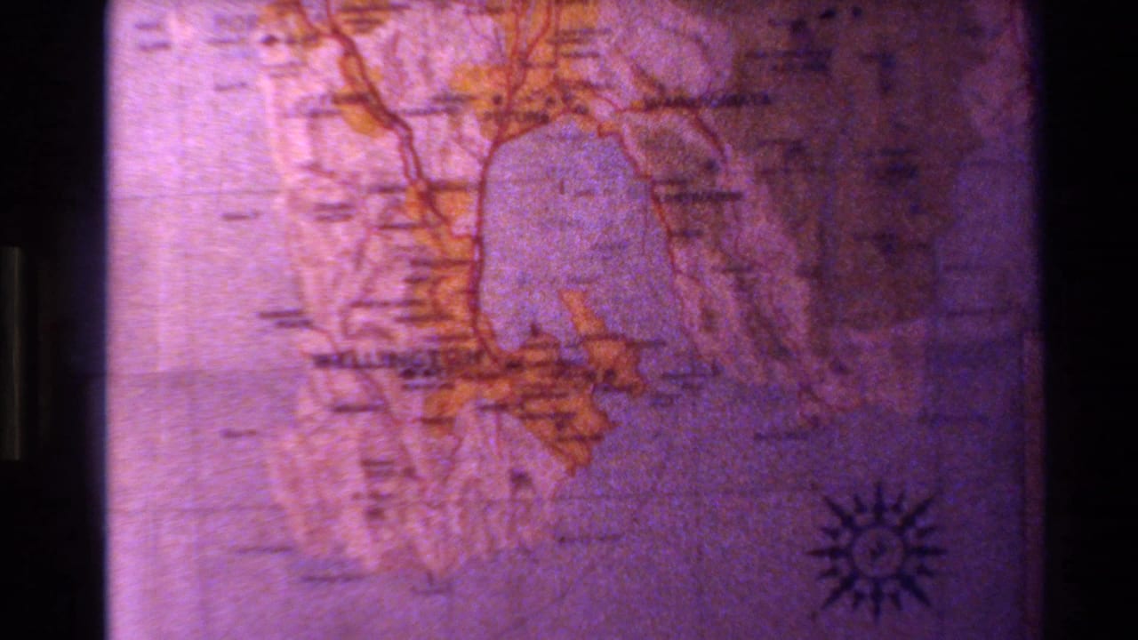 Projected map of New Zealand from 1981 home-movie footage, with orange geographic outlines, faint labels, and a compass rose visible.