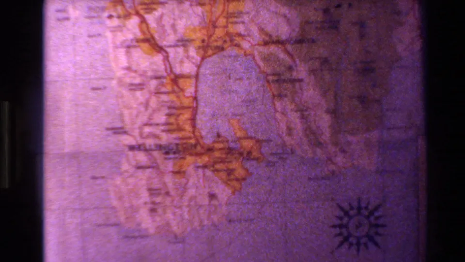 Projected map of New Zealand from 1981 home-movie footage, with orange geographic outlines, faint labels, and a compass rose visible.