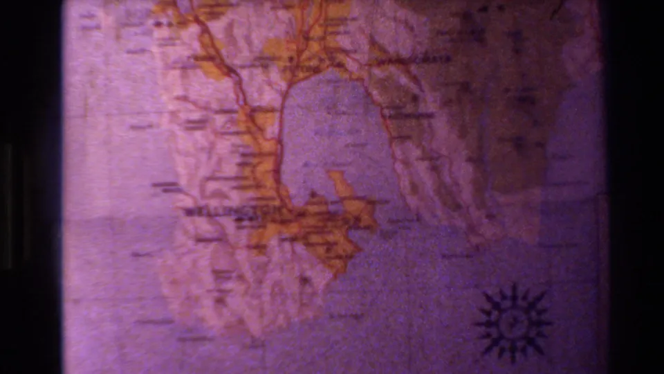 Soft 8mm projection of a New Zealand map, showing faint labeling, orange land shapes, grain, and dark projector framing.