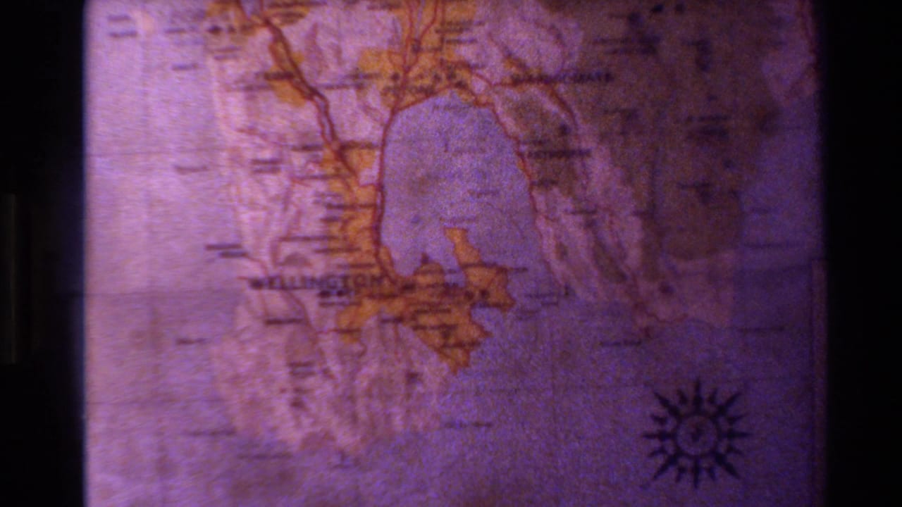 1981 New Zealand home-movie frame showing a close-up map detail with purple color cast, yellow land areas, and a compass rose.