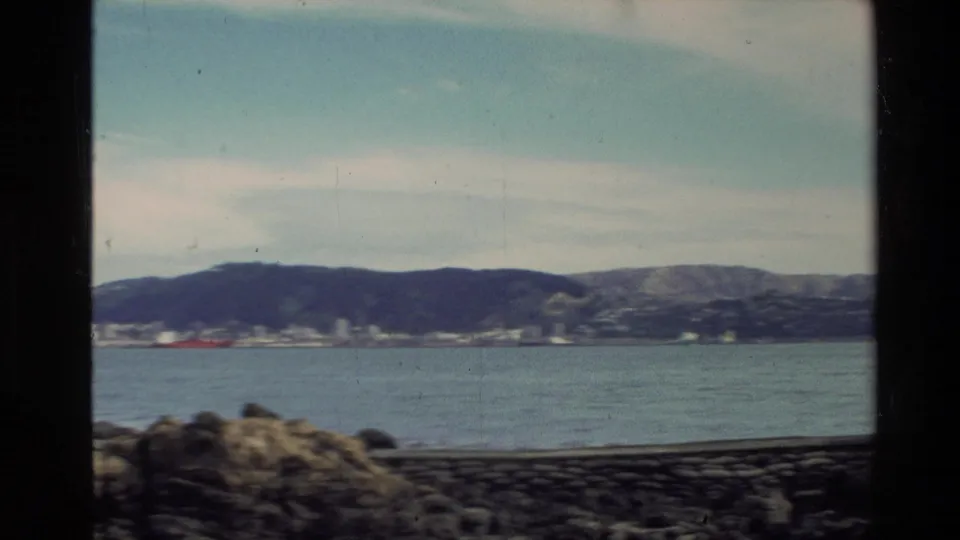 1981 New Zealand home-movie view across harbor water from a rocky shoreline toward distant hills and waterfront buildings.
