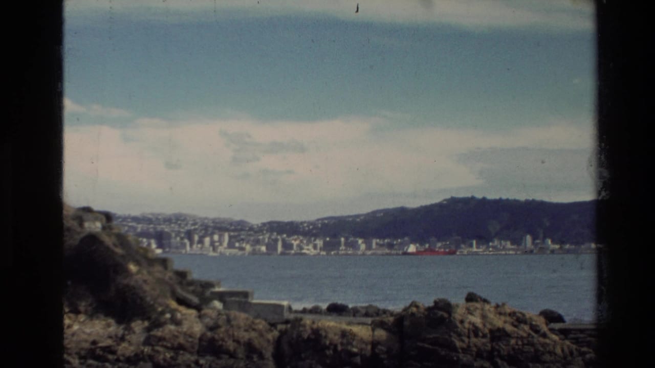 1981 New Zealand home-movie view of a coastal city across water, framed by dark film edges and rocky foreground.