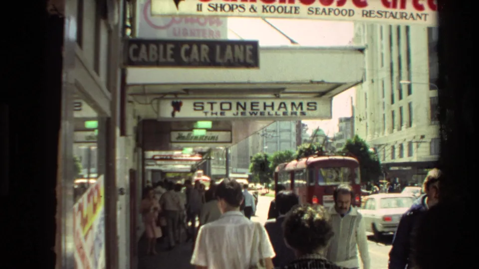 1981 New Zealand city sidewalk with pedestrians, Cable Car Lane signage, Stonehams sign, cars, and a red trolley in the street.