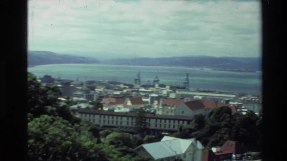 1981 New Zealand 8mm frame showing a coastal city panorama with harbor water, industrial cranes, rooftops, trees, and distant hills.