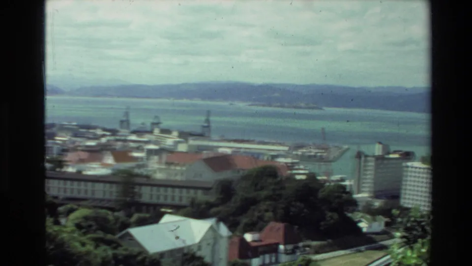 1981 New Zealand 8mm frame with a wide harbor view, city buildings below, cranes along the waterfront, and hills beyond the water.
