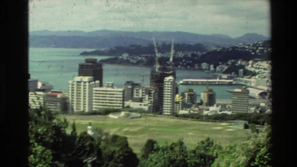 1981 New Zealand 8mm frame showing a harbor city, construction cranes, modern towers, green open land, bay water, and residential hills.