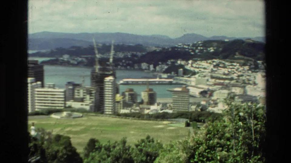 1981 New Zealand 8mm view from a hillside or park toward a coastal city, harbor, distant hills, and modern high-rise buildings.