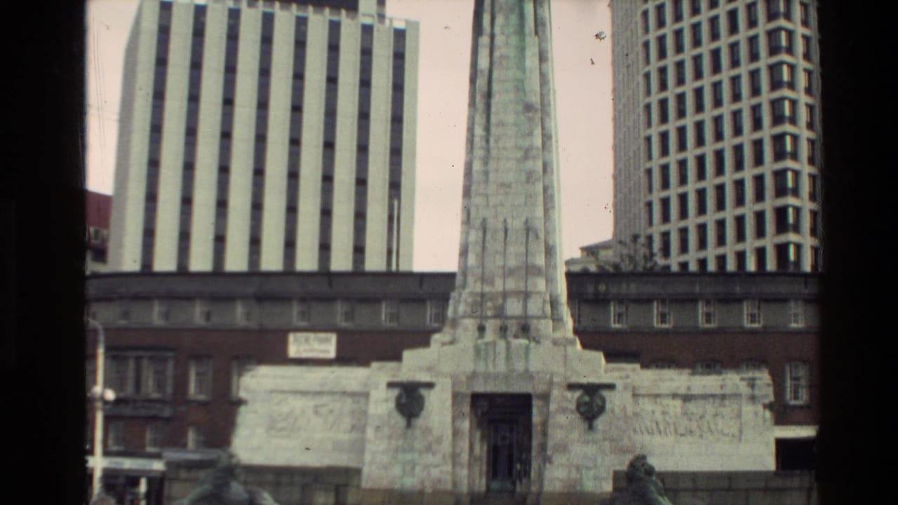 Street-level 8mm frame of a stone war memorial obelisk set against tall metropolitan office buildings in New Zealand.