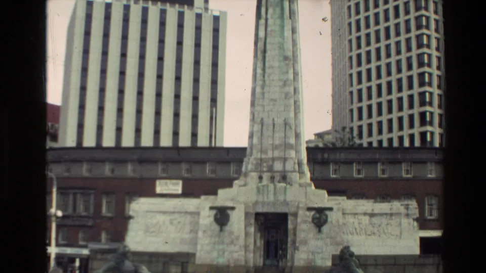 Street-level 8mm frame of a stone war memorial obelisk set against tall metropolitan office buildings in New Zealand.