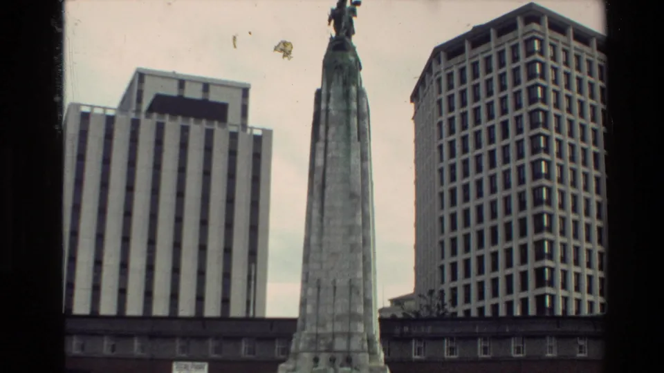 1981 New Zealand home-movie view looking up at a public memorial column with a statue, flanked by modern city office blocks.