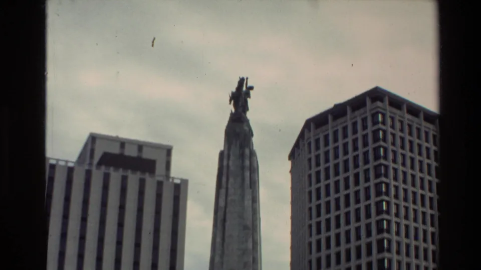 Close framing of the upper stone memorial and statue beside tall office buildings, consistent with 1981 New Zealand city footage.