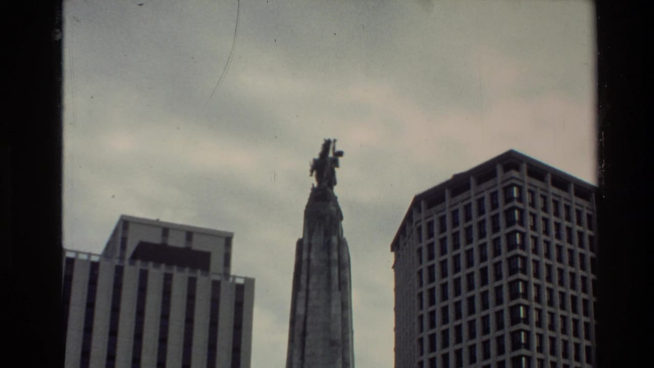 1981 New Zealand home-movie frame showing a tall monument flanked by modern city buildings and dark film gate edges.