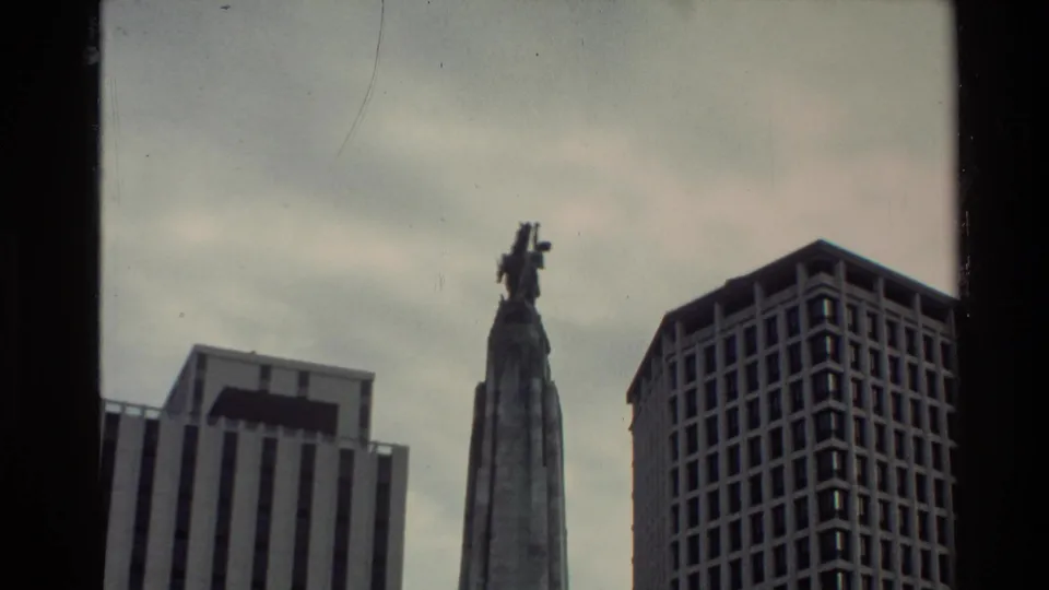 1981 New Zealand home-movie frame showing a tall monument flanked by modern city buildings and dark film gate edges.