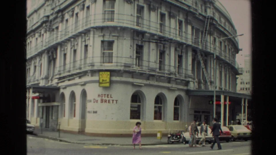 1981 New Zealand street scene showing the Hotel De Brett corner, pedestrians crossing or waiting, parked cars, and older commercial architecture.
