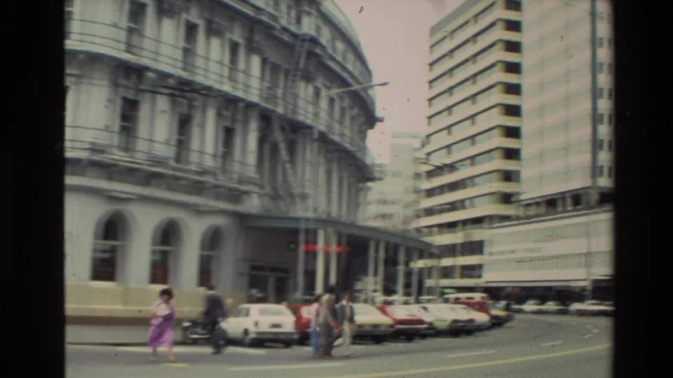New Zealand downtown frame from 1981 showing pedestrians near traffic lights, rows of cars, an older hotel facade, and a modern high-rise nearby.