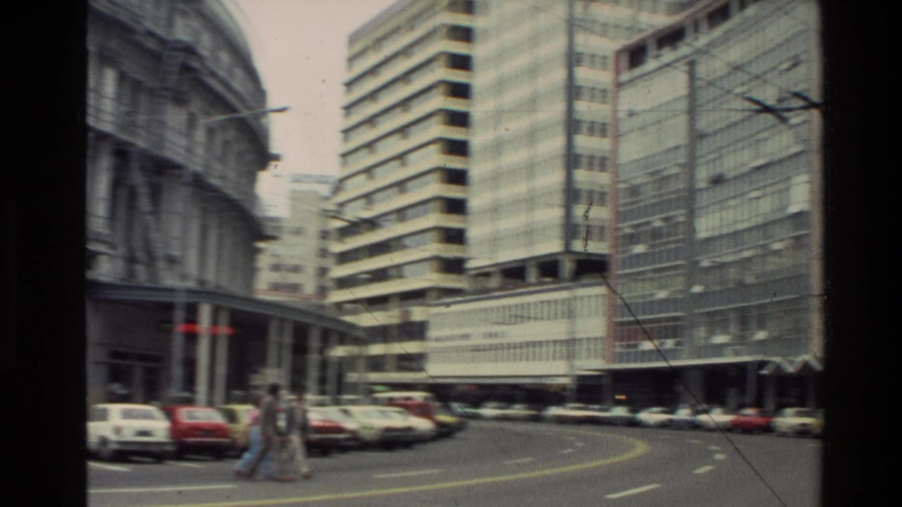 1981 New Zealand downtown street view with tall office buildings, parked cars, pedestrians, and overhead wires seen from a handheld camera.