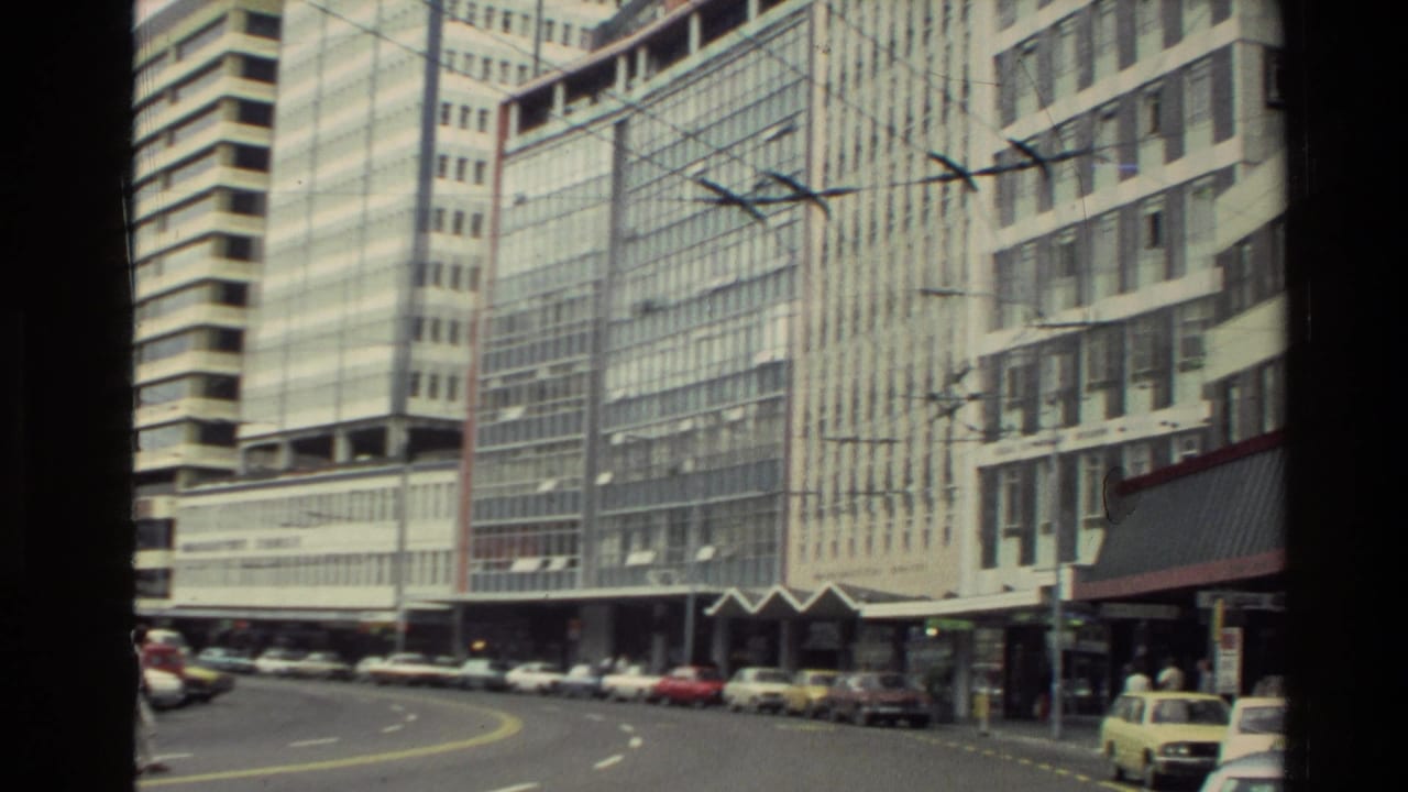 1981 New Zealand city street with glass-fronted office towers, parked cars, overhead wires, and a few pedestrians near storefronts.