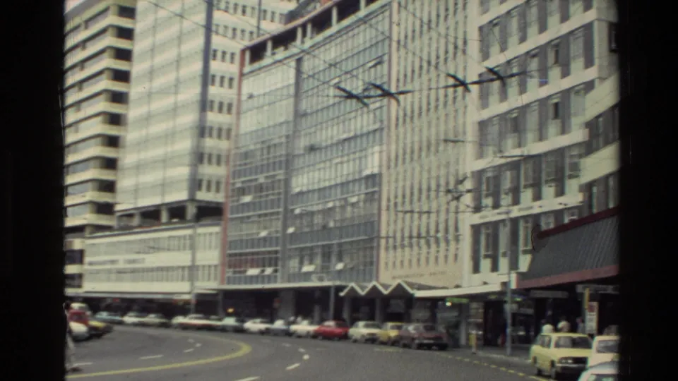 1981 New Zealand city street with glass-fronted office towers, parked cars, overhead wires, and a few pedestrians near storefronts.
