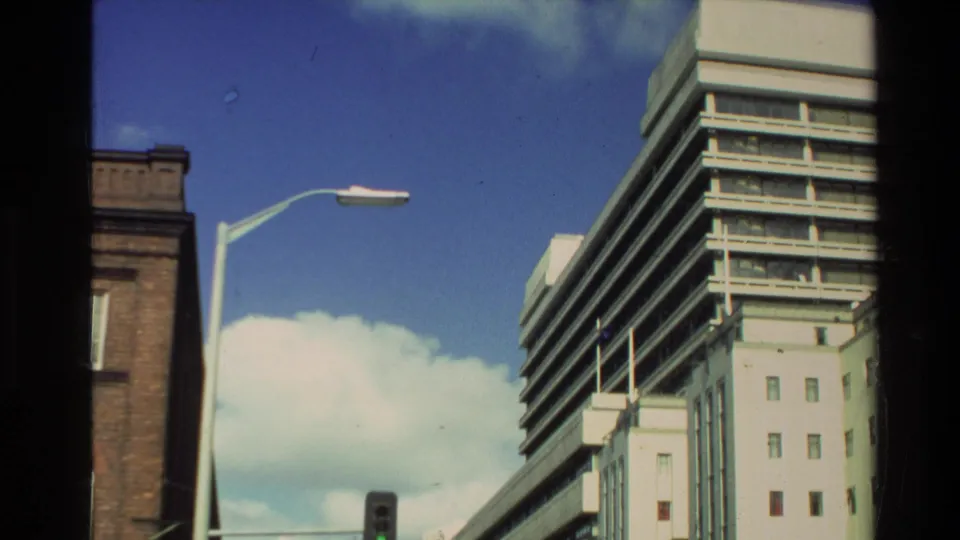 1981 New Zealand street view with a tall modern building, older brick facade, streetlamp, blue sky, and clouds.