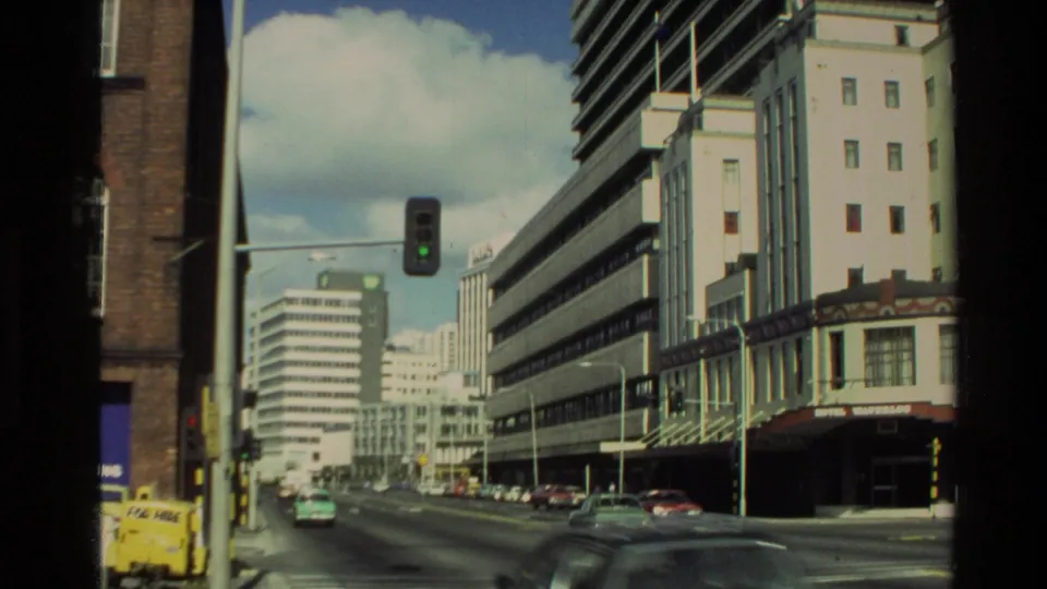 1981 New Zealand street scene with moving cars, a green traffic signal, tall office blocks, and visible storefront signage.