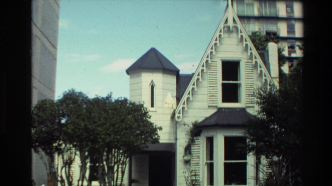 1981 New Zealand home-movie frame showing a white Victorian-style house with decorative gable trim, turret, trees, and modern buildings behind.