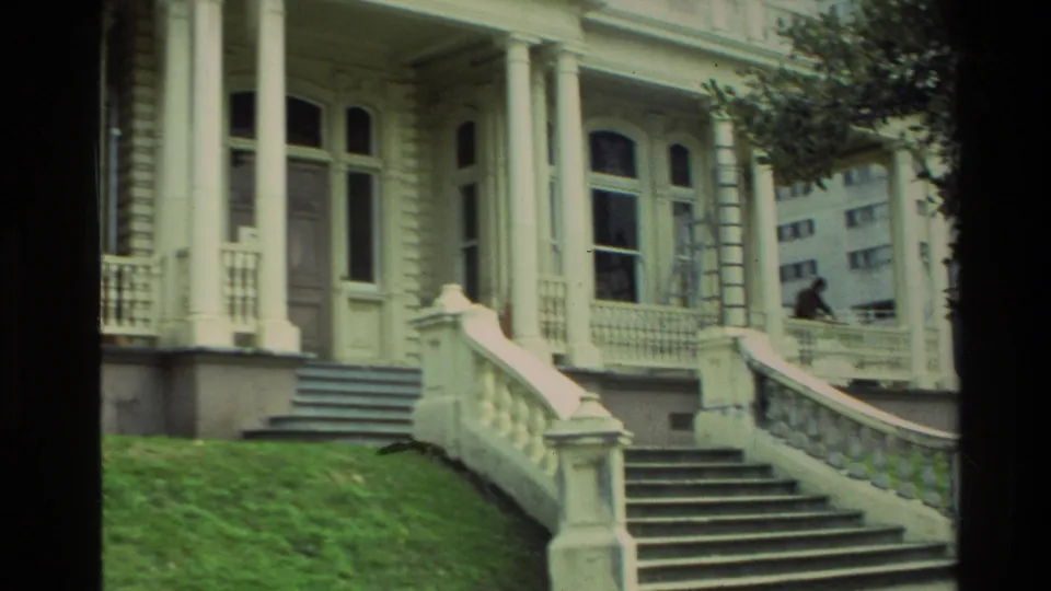 Street-level view of the ornate house porch with columns, tall windows, stairs, lawn, trees, and a briefly visible person near the railing.
