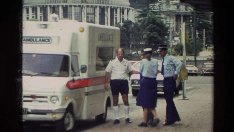 1981 New Zealand frame showing a man in a white shirt near an ambulance with two uniformed personnel on a city street.