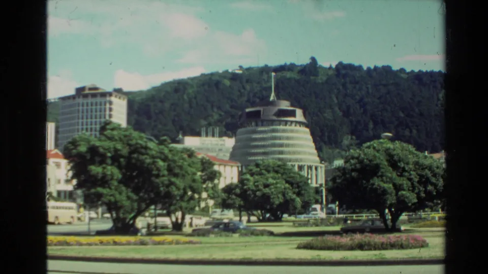 1981 New Zealand city view with civic buildings, roadside greenery, a landscaped area, and wooded hills beyond.