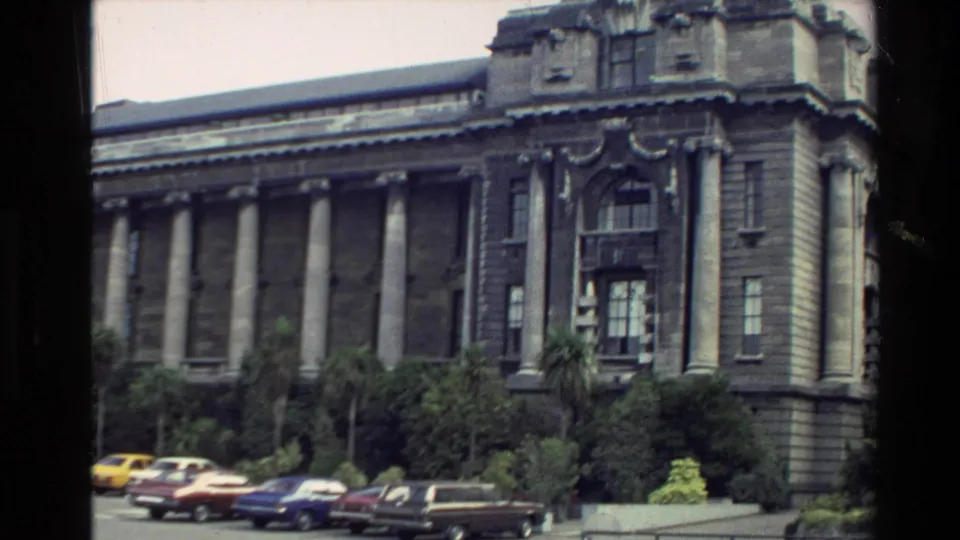 A columned civic building in New Zealand is filmed from the street, with parked cars and greenery in front.
