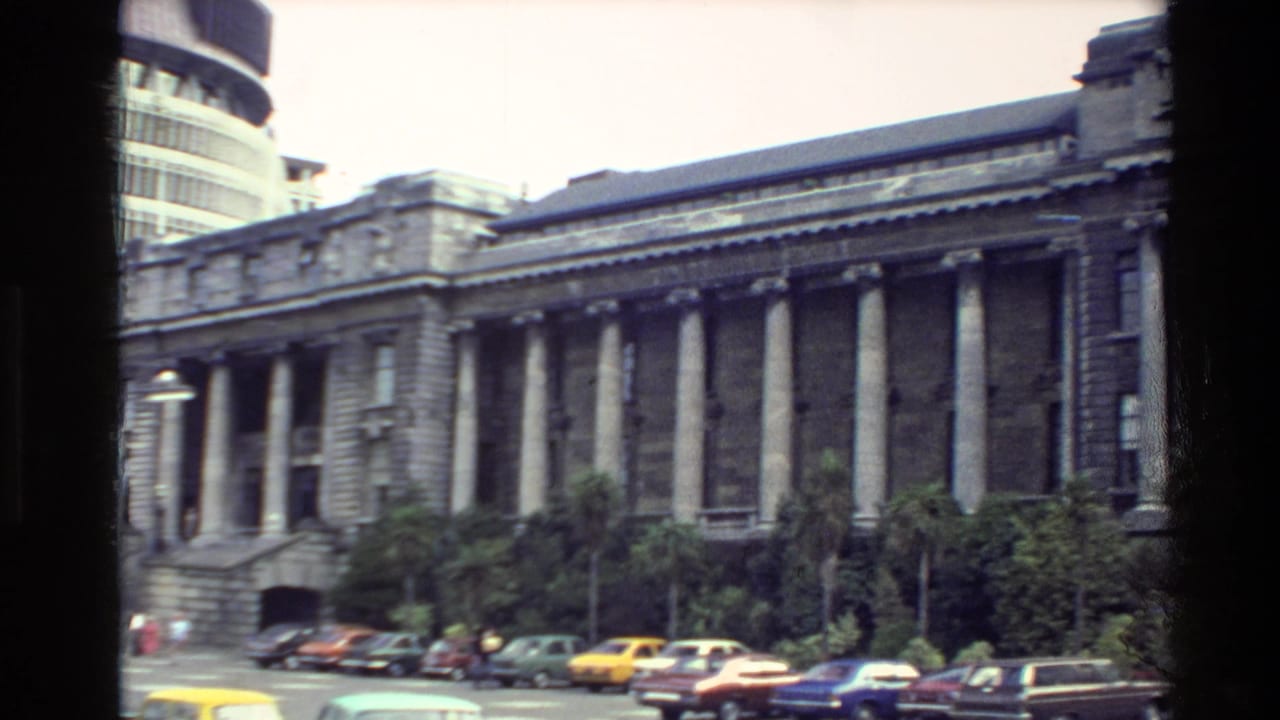 1981 New Zealand home-movie view of a classical stone government building, curbside cars, palm trees, and the edge of the Beehive.