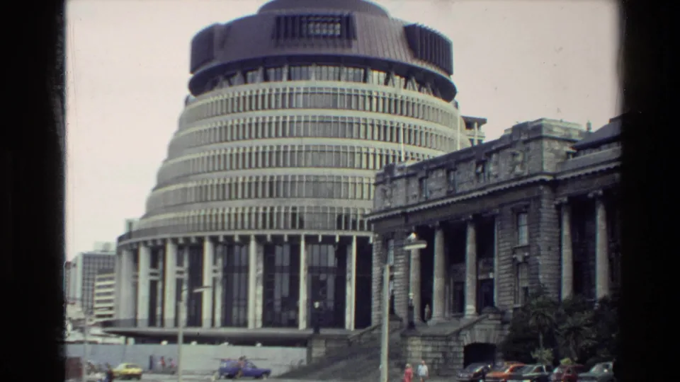 Street-level 1981 New Zealand view of the Beehive next to a classical facade, with colorful cars moving or parked in the foreground.
