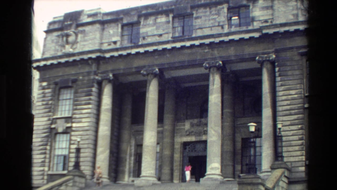 1981 New Zealand frame of a classical stone facade, showing columns, steps, and a person entering or standing at the doorway.