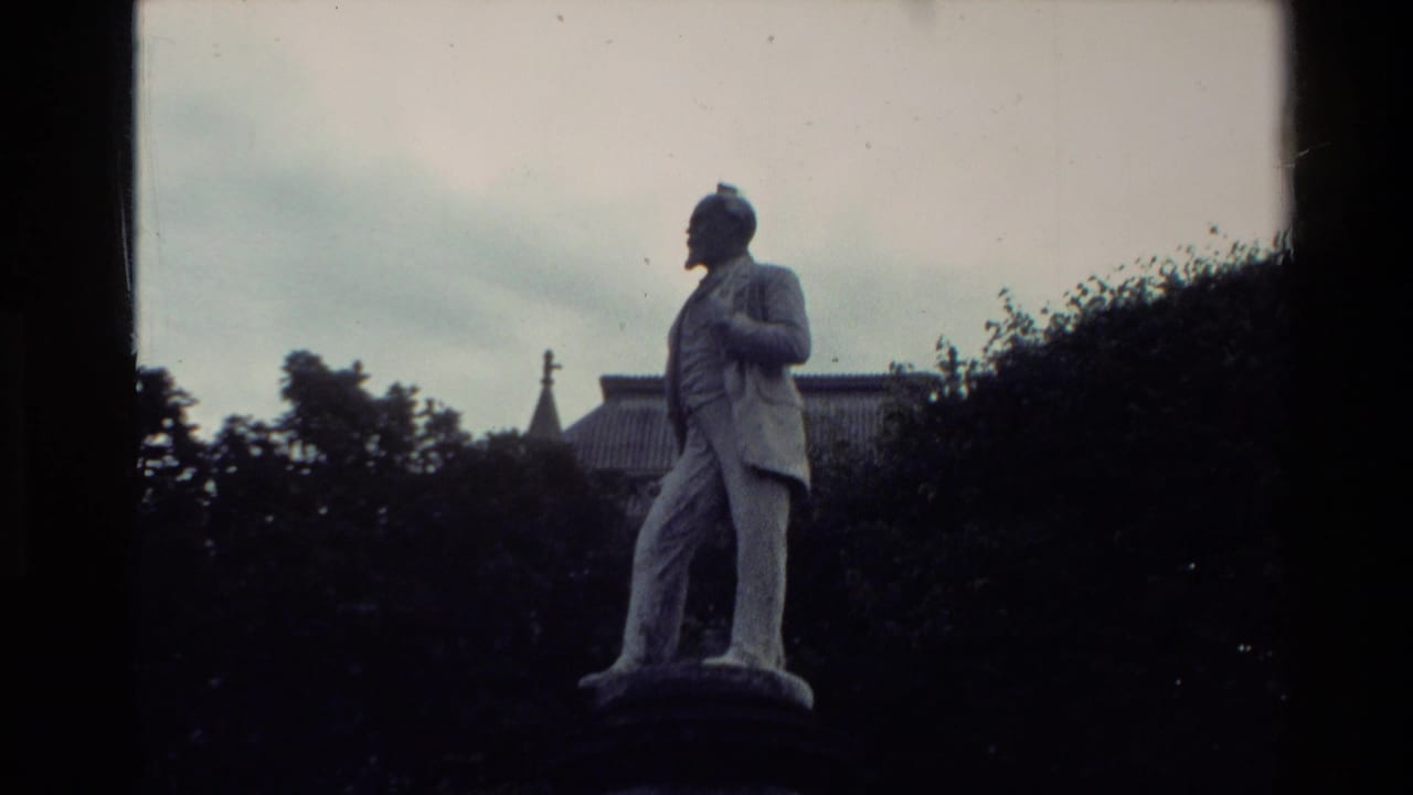 Full stone statue of a suited man standing outdoors with trees and rooflines behind it.