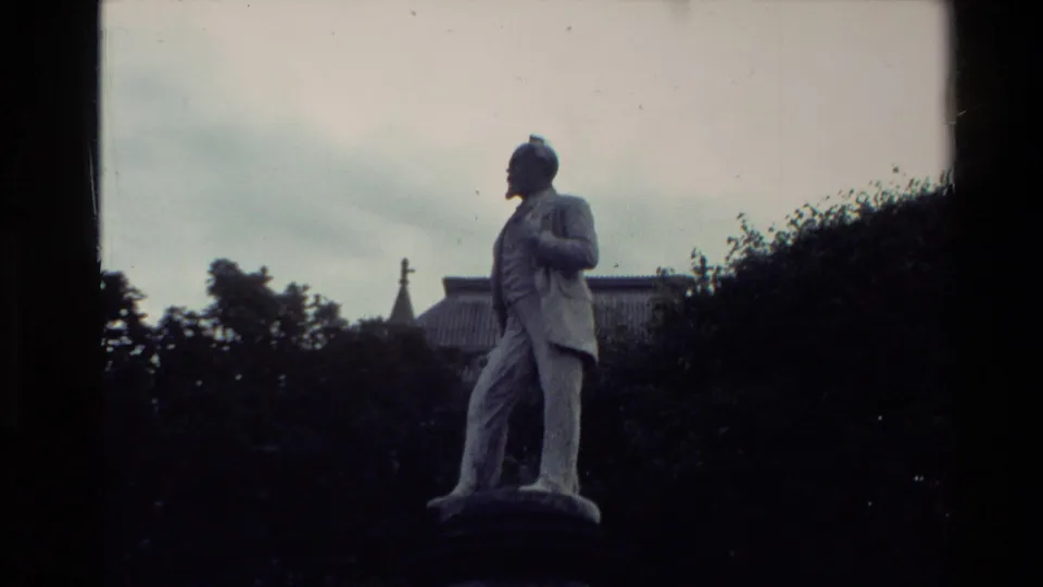 1981 New Zealand 8mm frame showing a full stone statue of a suited male figure in a park-like setting with trees and a building roof behind.