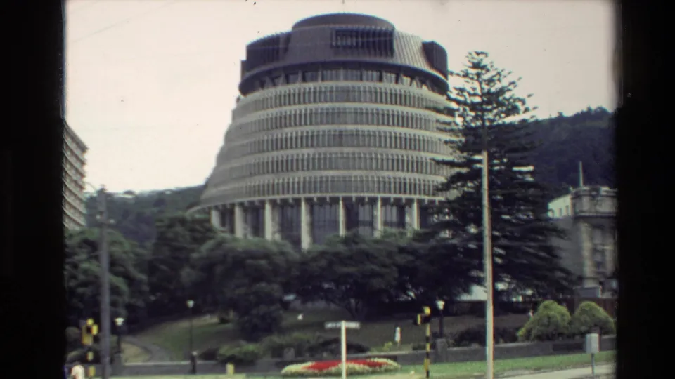 1981 New Zealand home-movie frame showing the Beehive building in Wellington with trees, road markings, and landscaped public grounds.