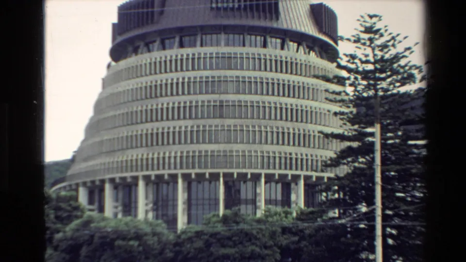 1981 New Zealand frame showing Wellington's circular Beehive building framed by trees, a streetlight, and low urban greenery.