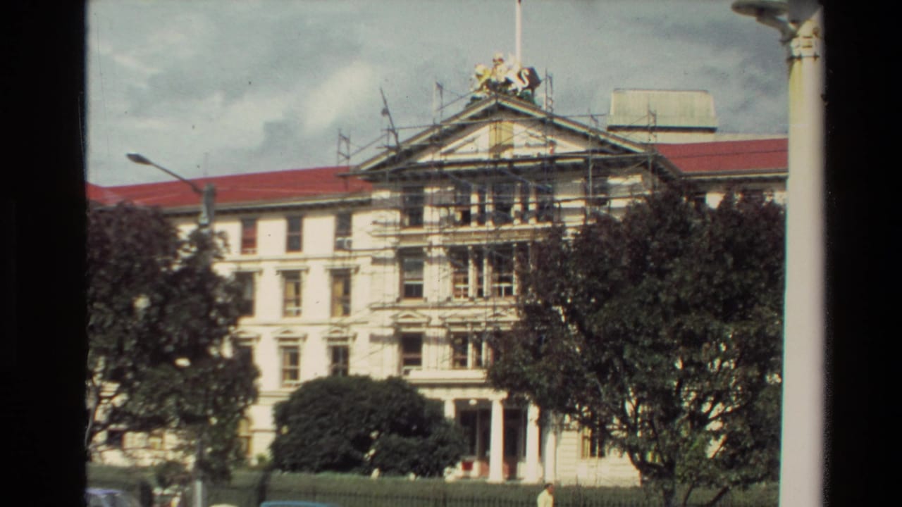 1981 New Zealand exterior view of a classical-style building with a red roof, front scaffolding, trees, and a roof crest or ornament above the pediment.