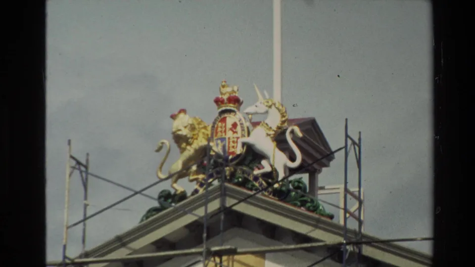 Close 1981 New Zealand frame of a royal coat of arms with a golden lion, white unicorn, shield, crown, flagpole, and scaffolding on a roofline.